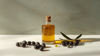 Editorial still-life of a glass bottle of golden olive oil with olives, an olive branch sprig and a dropper, styled on cream and stone in deep olive, dark brown and lime-green tones, no text