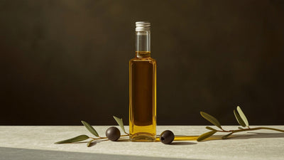 Editorial still-life of a glass bottle of fresh-pressed golden olive oil with whole olives and an olive branch sprig, styled on cream and stone in deep olive-green and warm brown tones.