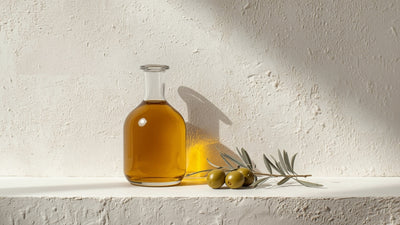Editorial still-life of a glass bottle of golden olive oil with olives and an olive branch sprig on textured cream stone, in Oliiv's dark olive, lime-accent palette, no text